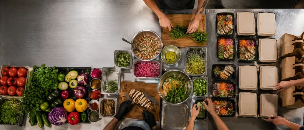 Overhead view of a stainless prep table with produce, pans and packed containers