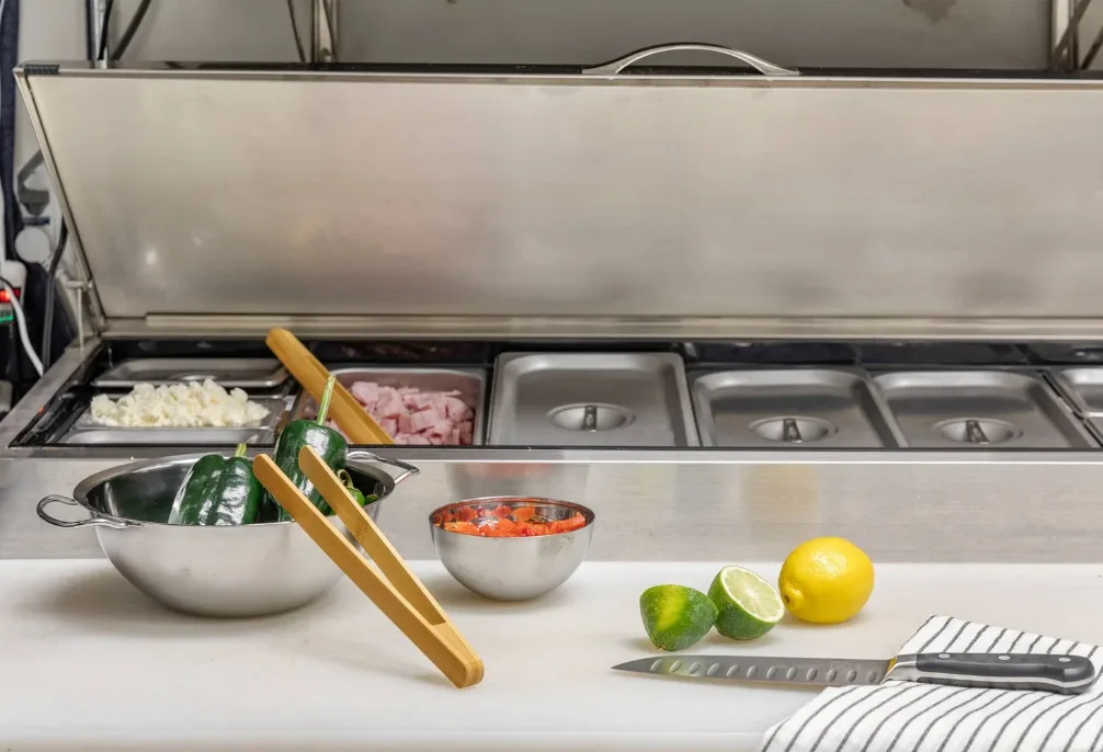 Stainless refrigerated prep rail with pans, alongside a clean cutting surface